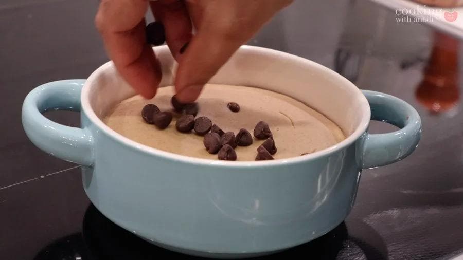 Hand sprinkling chocolate chips onto baked oats in a light blue ramekin with a creamy center.