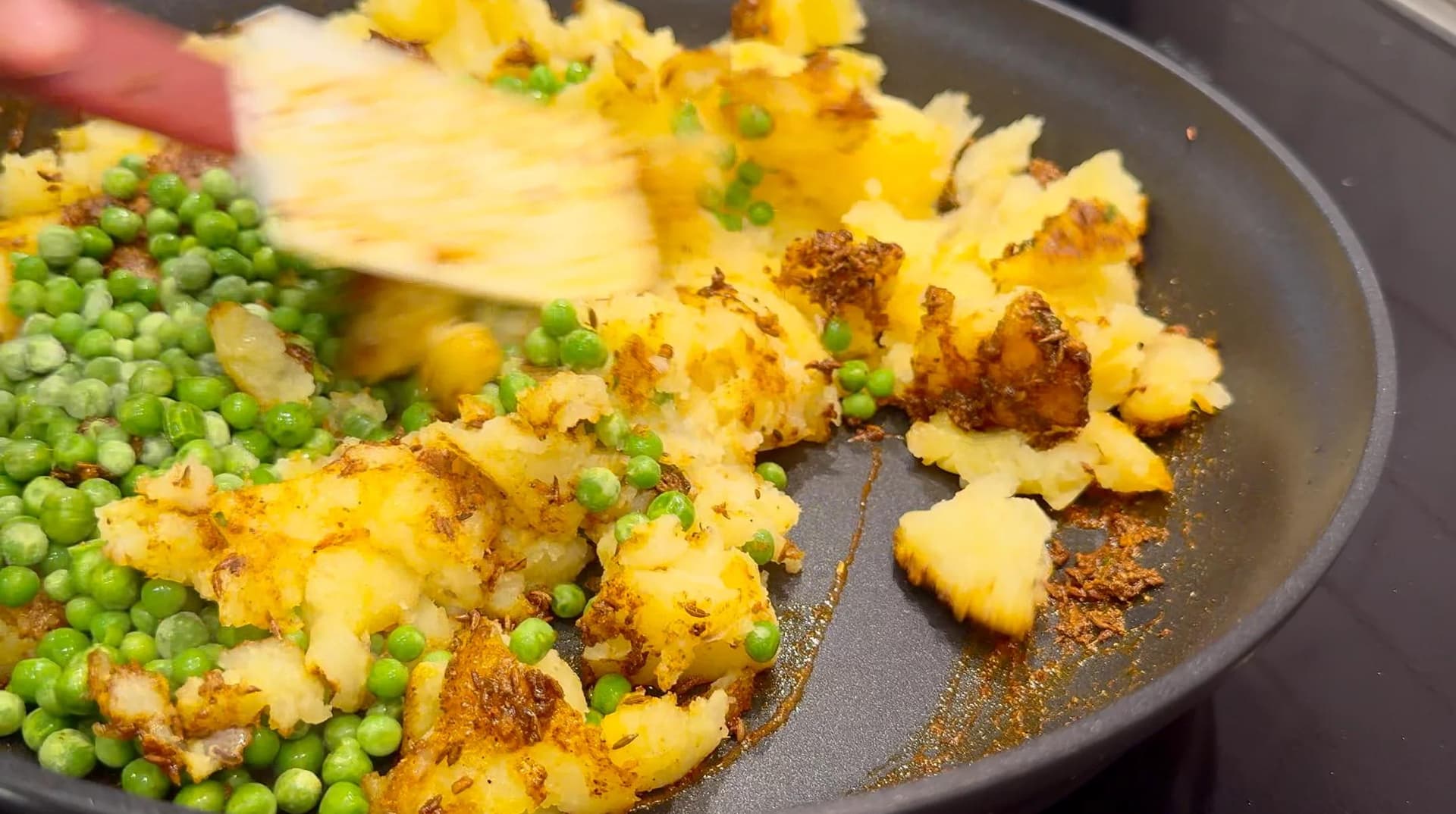 Mashed potatoes and peas being stirred together in a dark pan with roasted spices and a spatula.