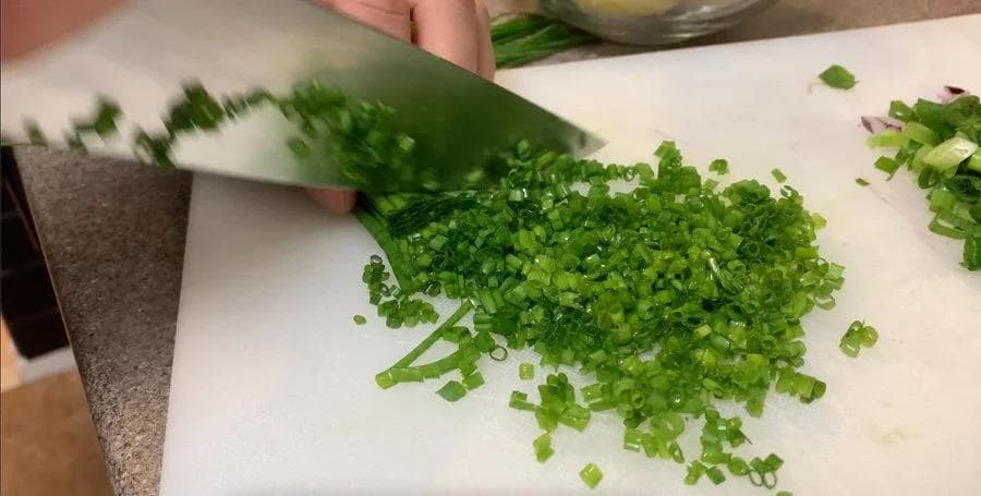 Finely chopped green onions and chives on a cutting board with a knife.
