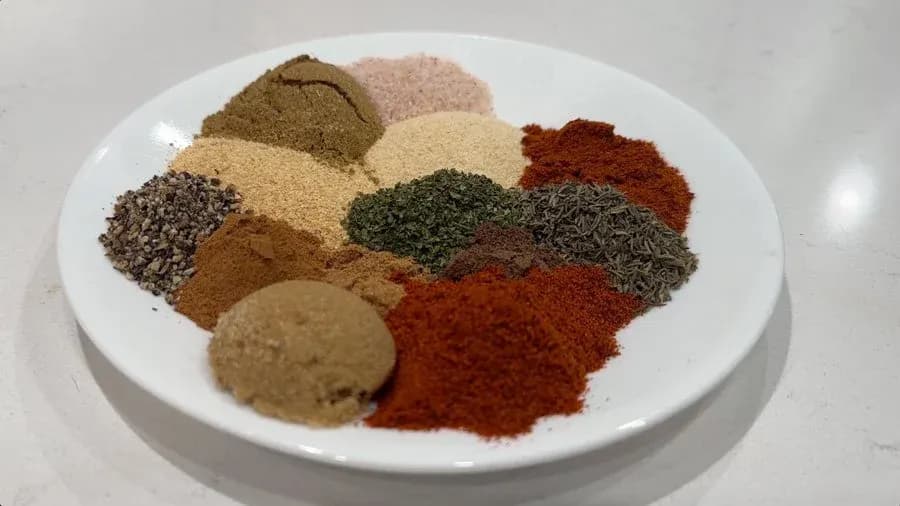 Individual jerk marinade spices arranged on a white plate, including cumin seeds, garlic powder, paprika, and dried herbs.