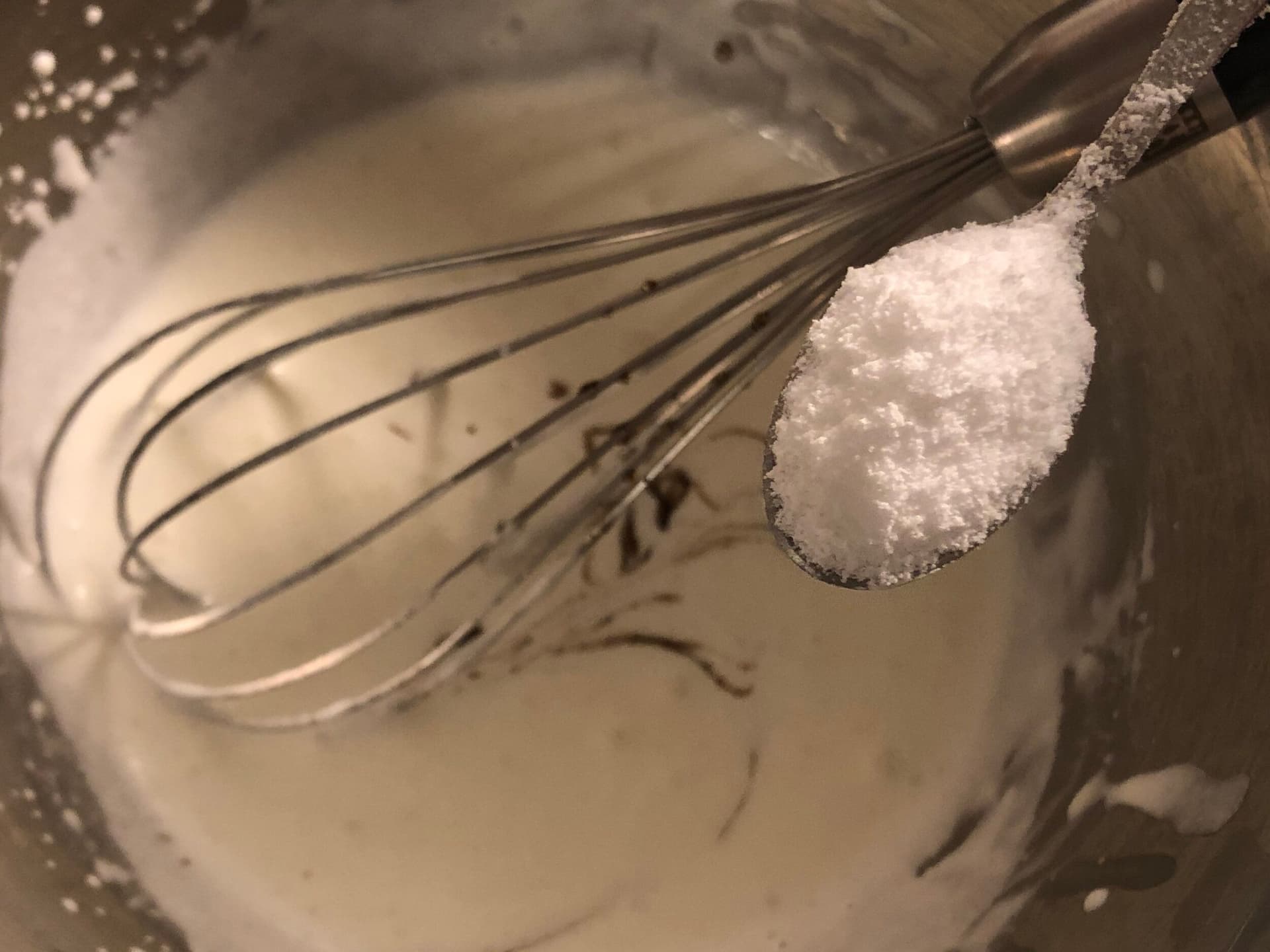 Whisk lifting soft peaks of freshly whipped cream with sugar on spoon held above bowl.