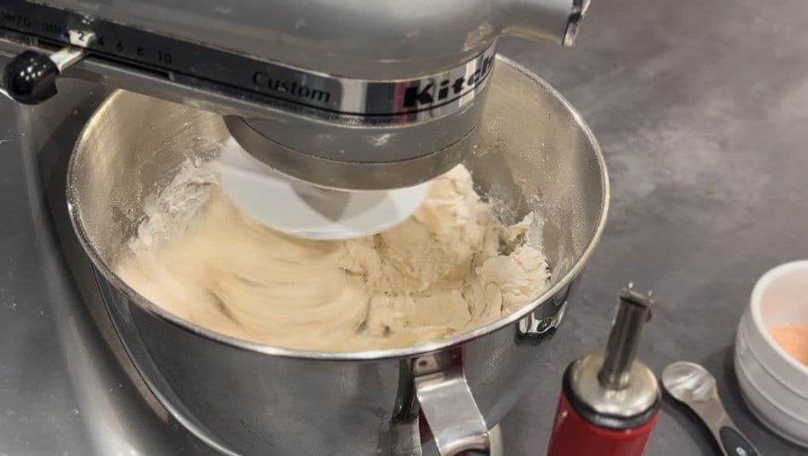 Kitchen Aid mixer with paddle attachment kneading smooth pizza dough in metal bowl during mixing process.