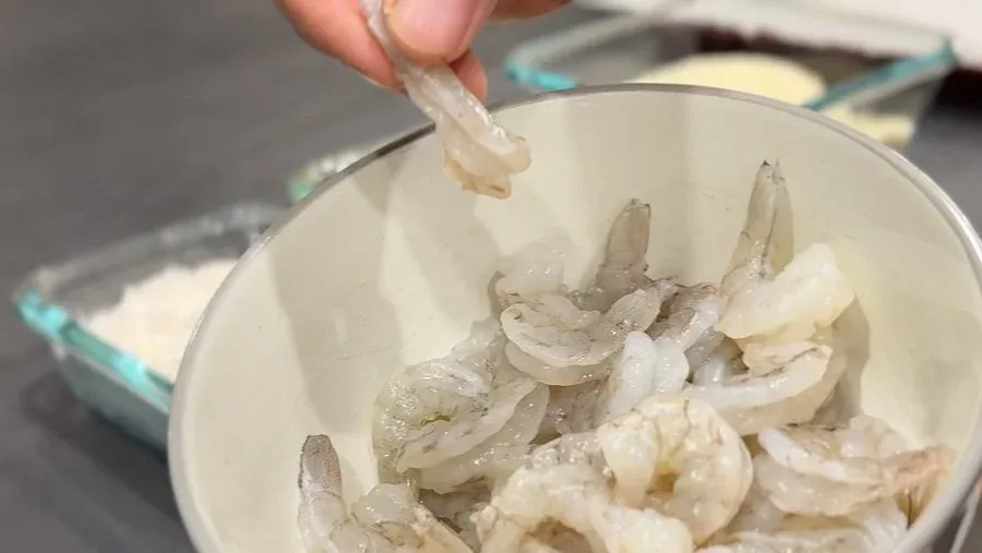 Hand dipping breaded shrimp into a white bowl during the coating process for popcorn shrimp.