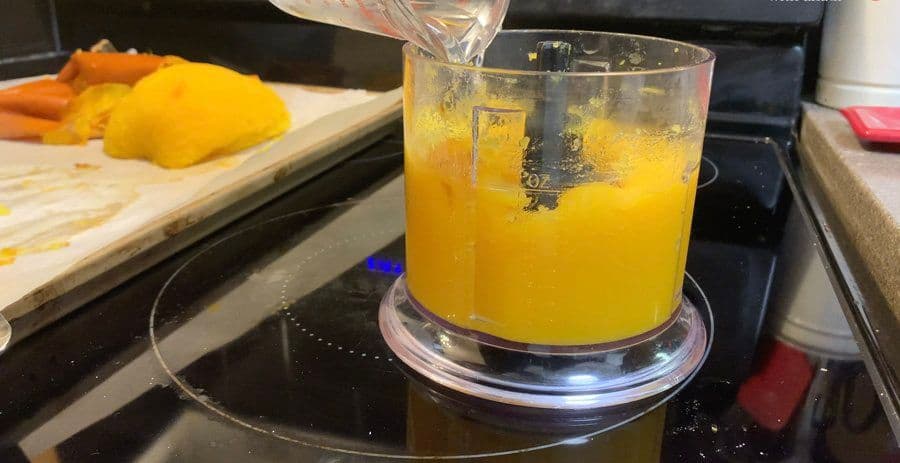 Food processor blending orange pumpkin puree with water on stovetop, fresh pumpkin visible on cutting board nearby.