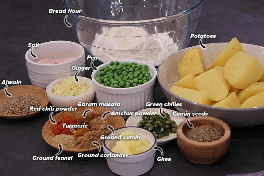 Ingredients for air fryer potato samosas displayed on a dark surface, including potatoes, peas, spices, flour, and aromatics.