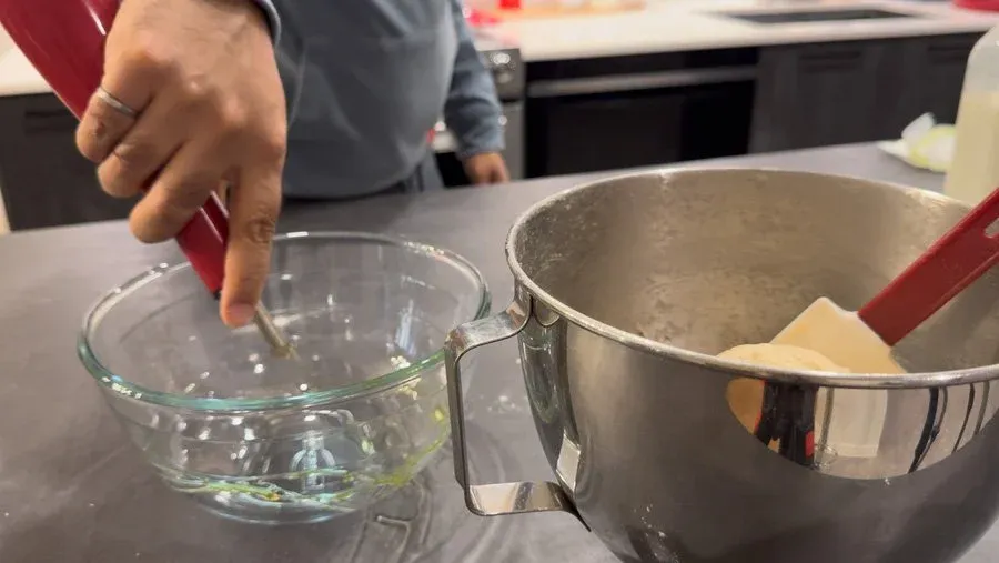 Hands brushing olive oil into a glass bowl next to a metal bowl containing dough during dinner roll preparation.