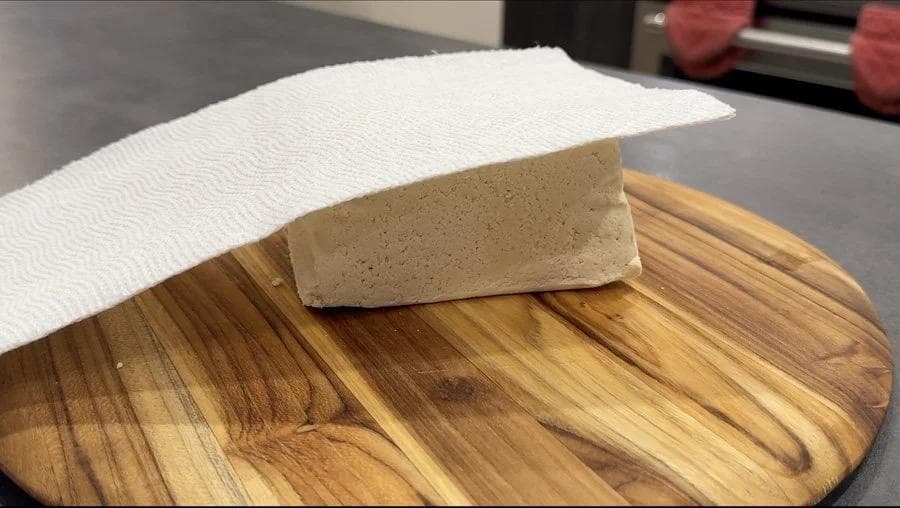 Block of tofu on wooden cutting board being pressed with paper towel to remove moisture.