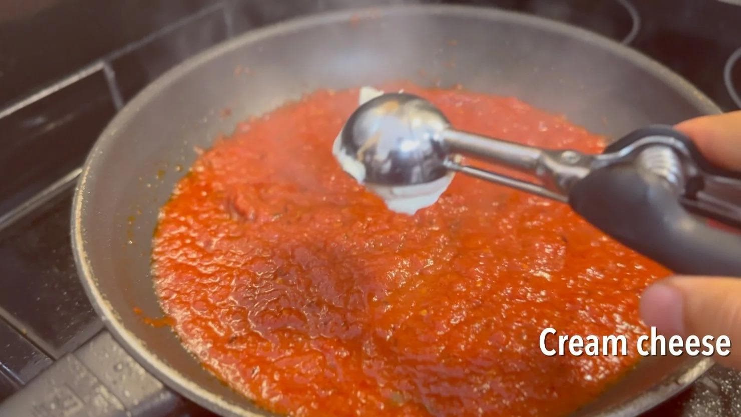 Cream cheese being added to a bubbling red tomato sauce in a pan with a spoon.