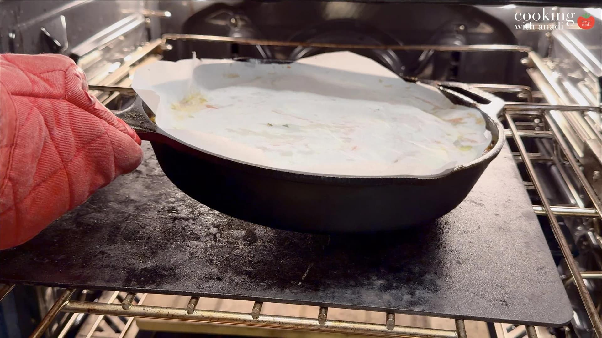 Cast iron skillet with cream-covered ratatouille being placed into oven, held with red oven mitt.