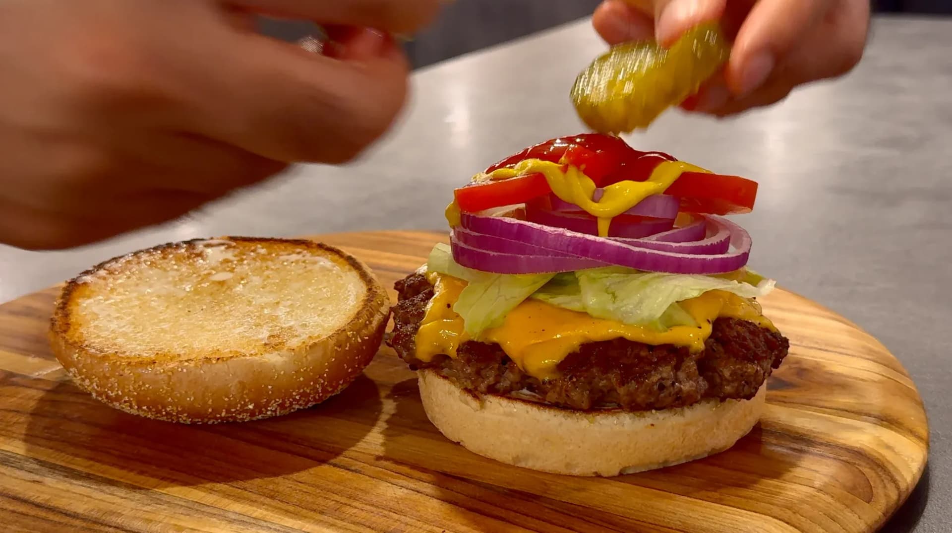 Classic cheeseburger on toasted bun with lettuce, cheese, red onion, tomato, and yellow pepper, with pickle being added on top.