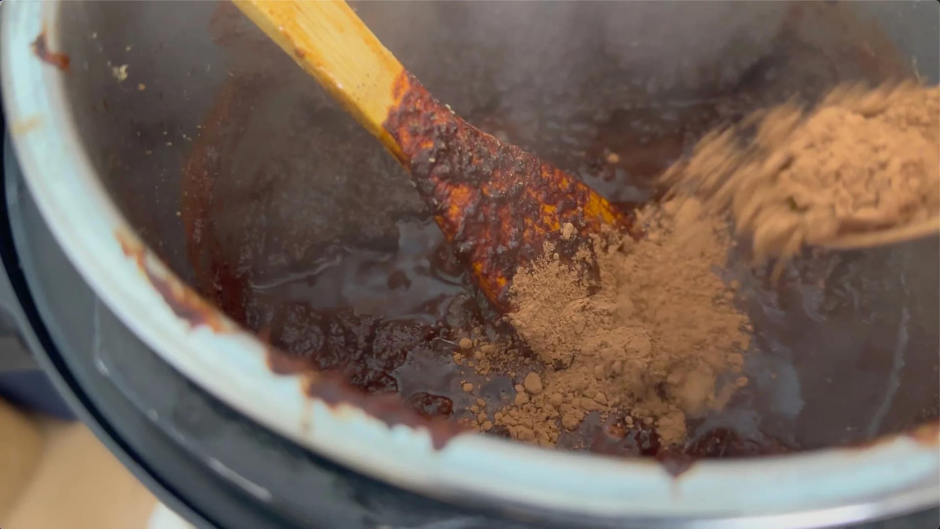 Wooden spoon stirring cocoa powder and ground spices into dark broth in a white bowl for chili preparation.