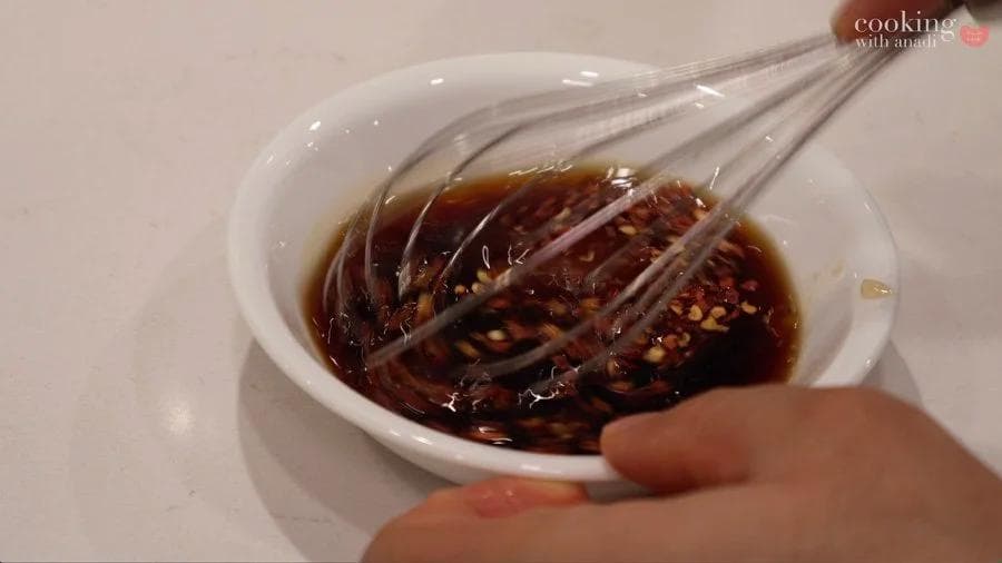 Whisking stir fry sauce with soy sauce, vinegar, and chili flakes in a white bowl with a whisk.