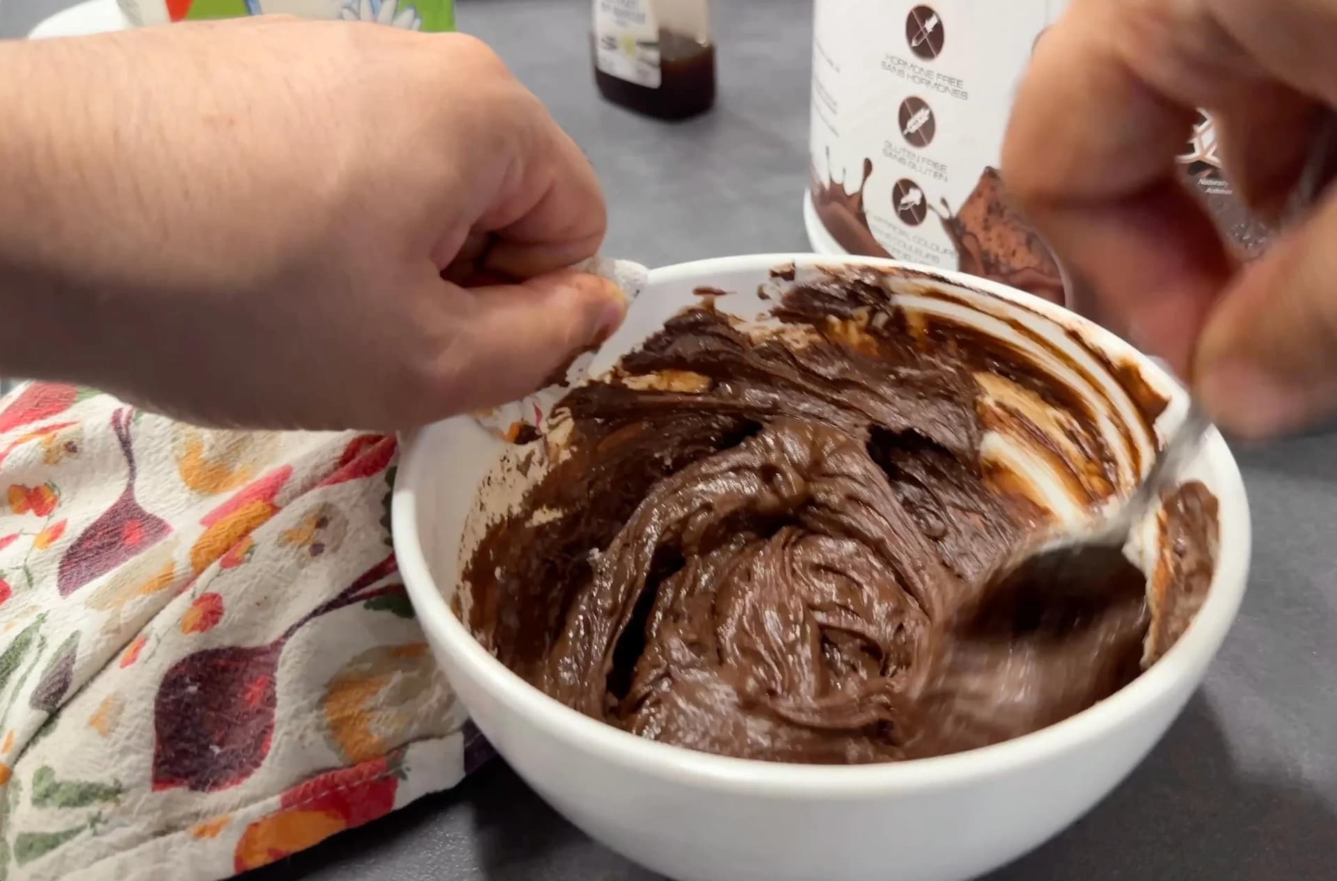 Hands stirring smooth, thick chocolate protein mixture in a white bowl with a spatula.