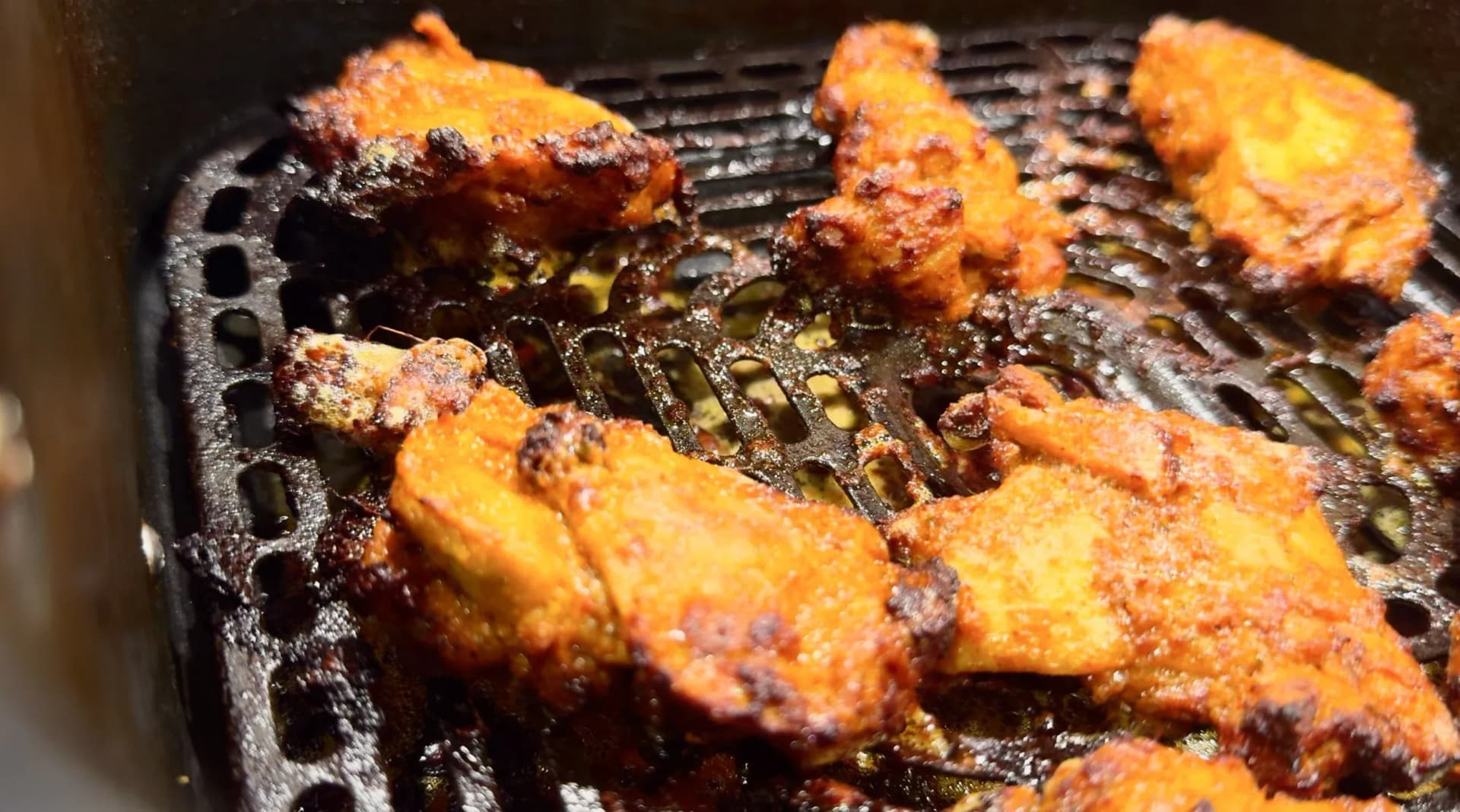 Golden-brown chicken tikka pieces with charred edges cooking in a black air fryer basket, glistening with butter baste.