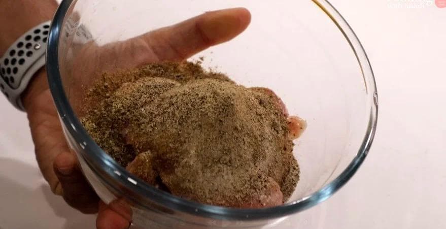 Hand holding glass bowl with chicken thigh coated in dry spice rub of oregano, cumin, and black pepper.