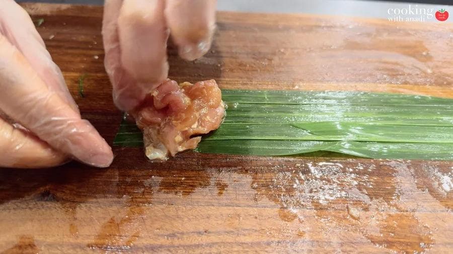Hands placing a piece of raw marinated chicken onto flat green pandan leaves on a wooden cutting board.