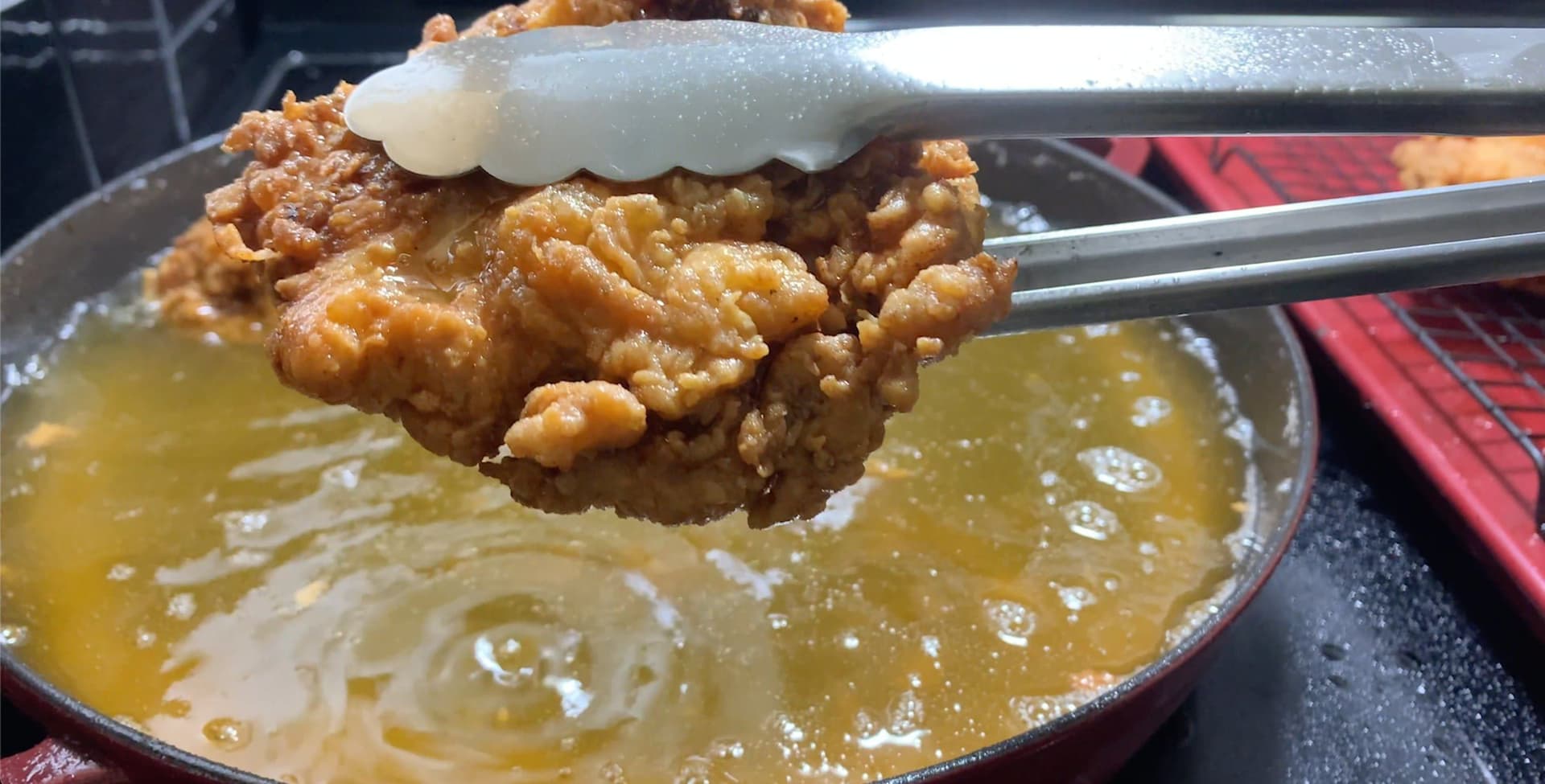 Crispy fried chicken piece being lifted from hot oil with chopsticks, topped with white garnish.