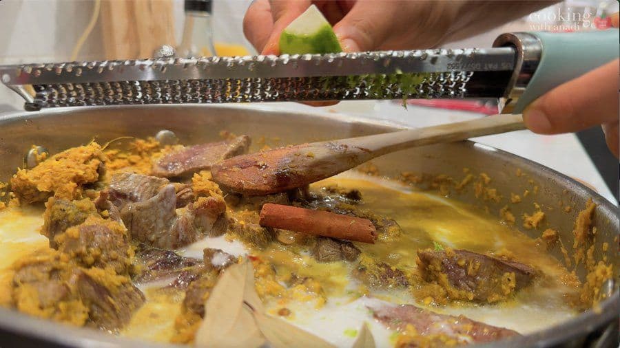 Beef rendang simmering in creamy golden sauce with cinnamon stick and bay leaves in a pan, being stirred with a wooden spoon.