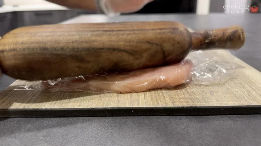 Wooden rolling pin flattening chicken breast wrapped in plastic wrap on a cutting board.