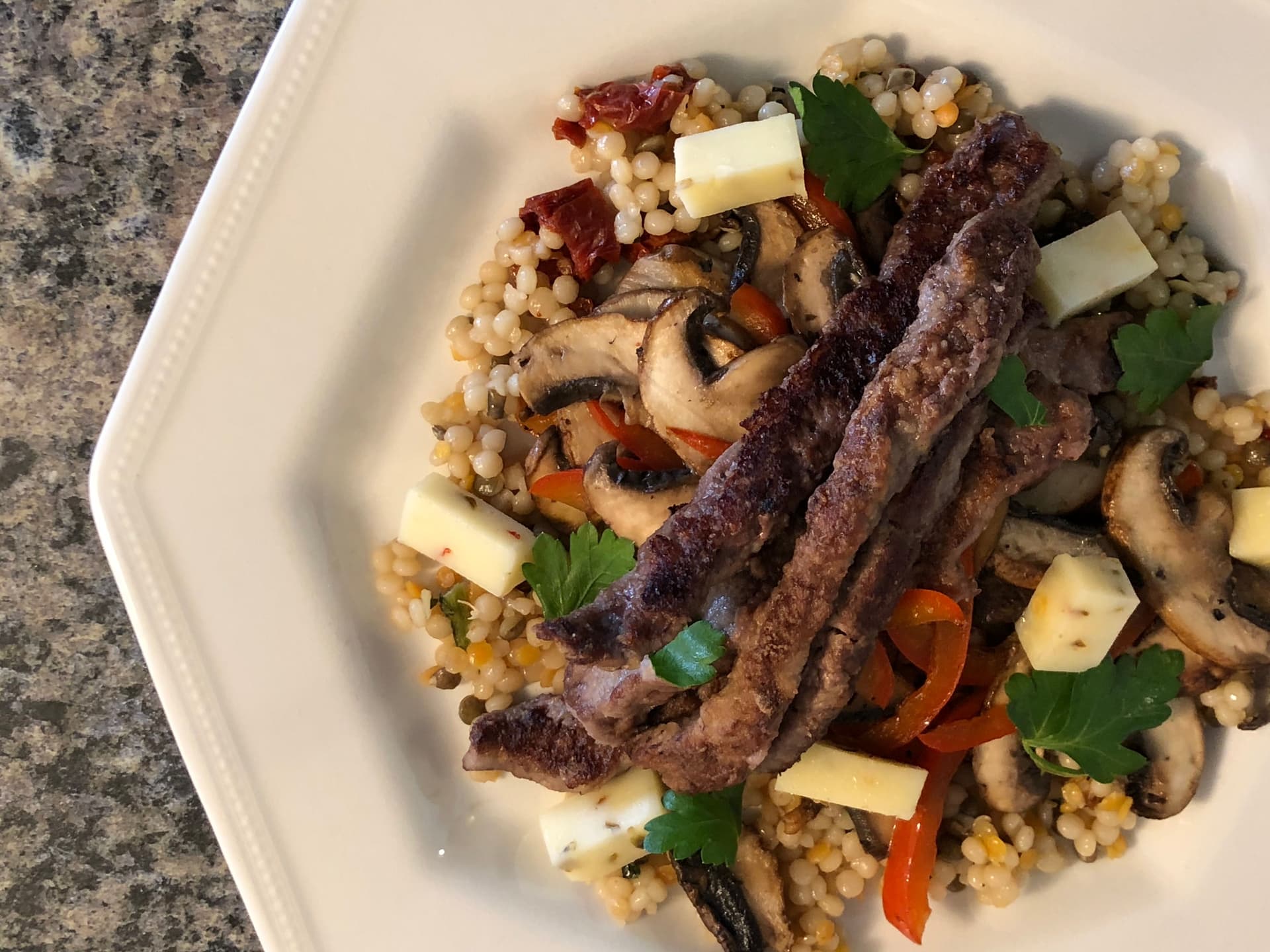 Sliced beef steak over couscous with mushrooms, red peppers, cheese cubes, and fresh parsley garnish.