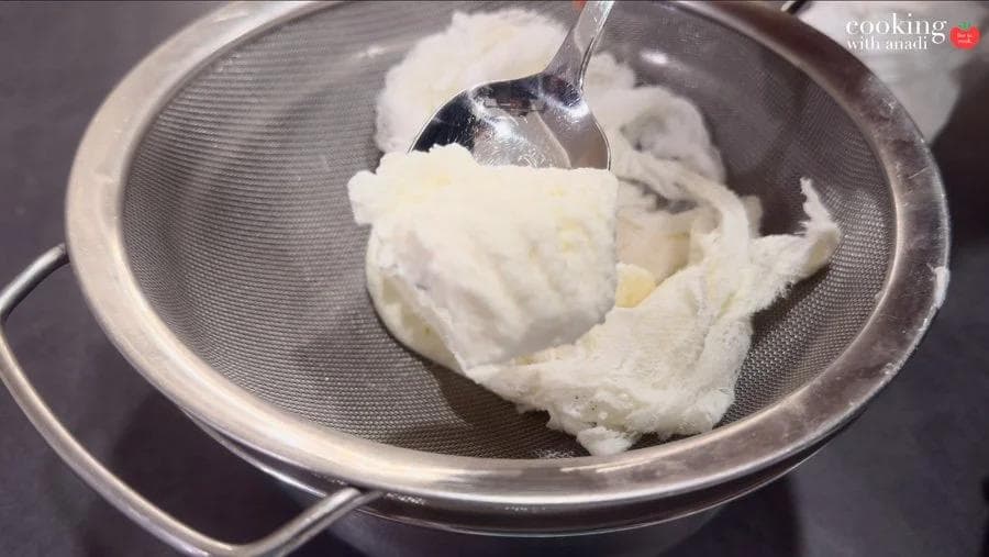 Thick hung curd being scooped from a fine mesh sieve strainer with a metal spoon after draining