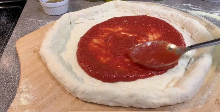 Pizza dough with raised crust edges on wooden board, red tomato sauce being spread with spoon.