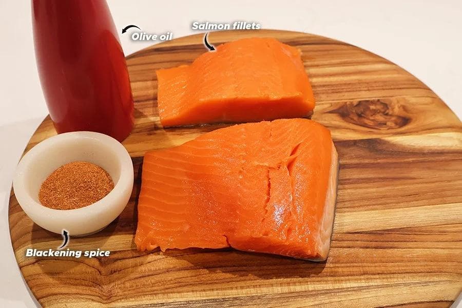 Salmon fillets, olive oil bottle, and blackening spice in a small bowl on a wooden cutting board.