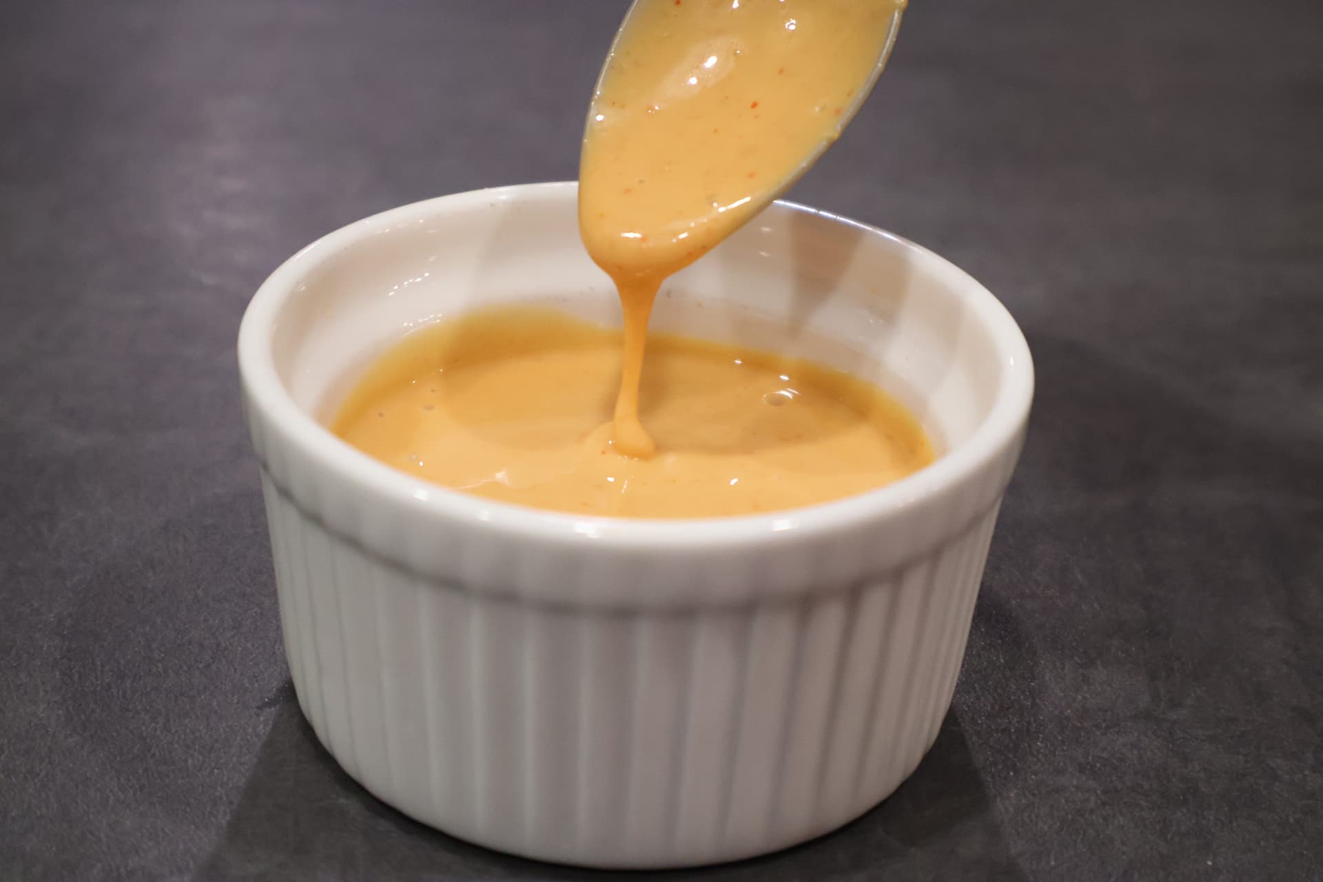 Spicy umami sauce being drizzled from a spoon into a white ramekin bowl on a dark surface.