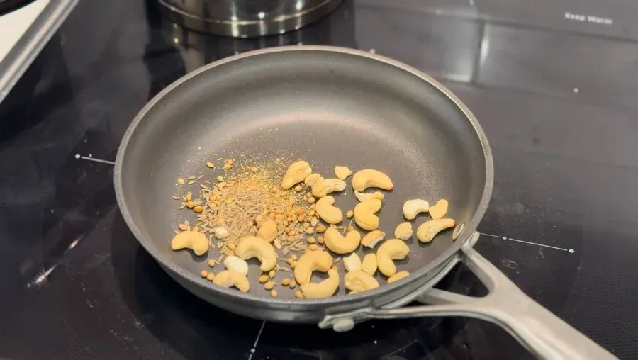 Cashews, cumin seeds, and coriander seeds being roasted in a gray pan on a stovetop until lightly browned.