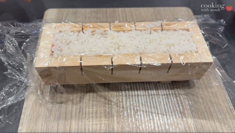 Wooden sushi mold filled with rice and wrapped in plastic, ready for pressing.