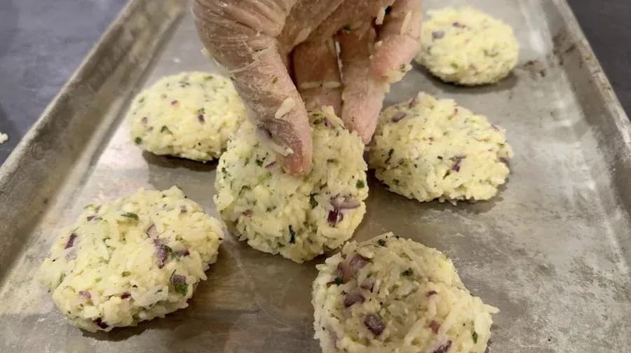 Hand shaping herb and cheese-filled rice fritter patties on a parchment-lined baking tray.