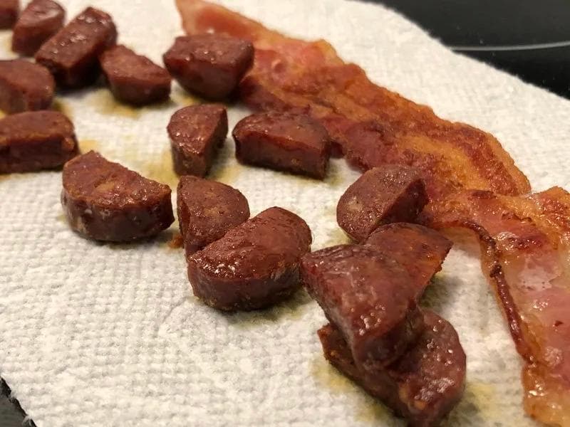 Cooked crispy bacon strips and diced chorizo pieces on a white paper towel with oil pooling underneath.