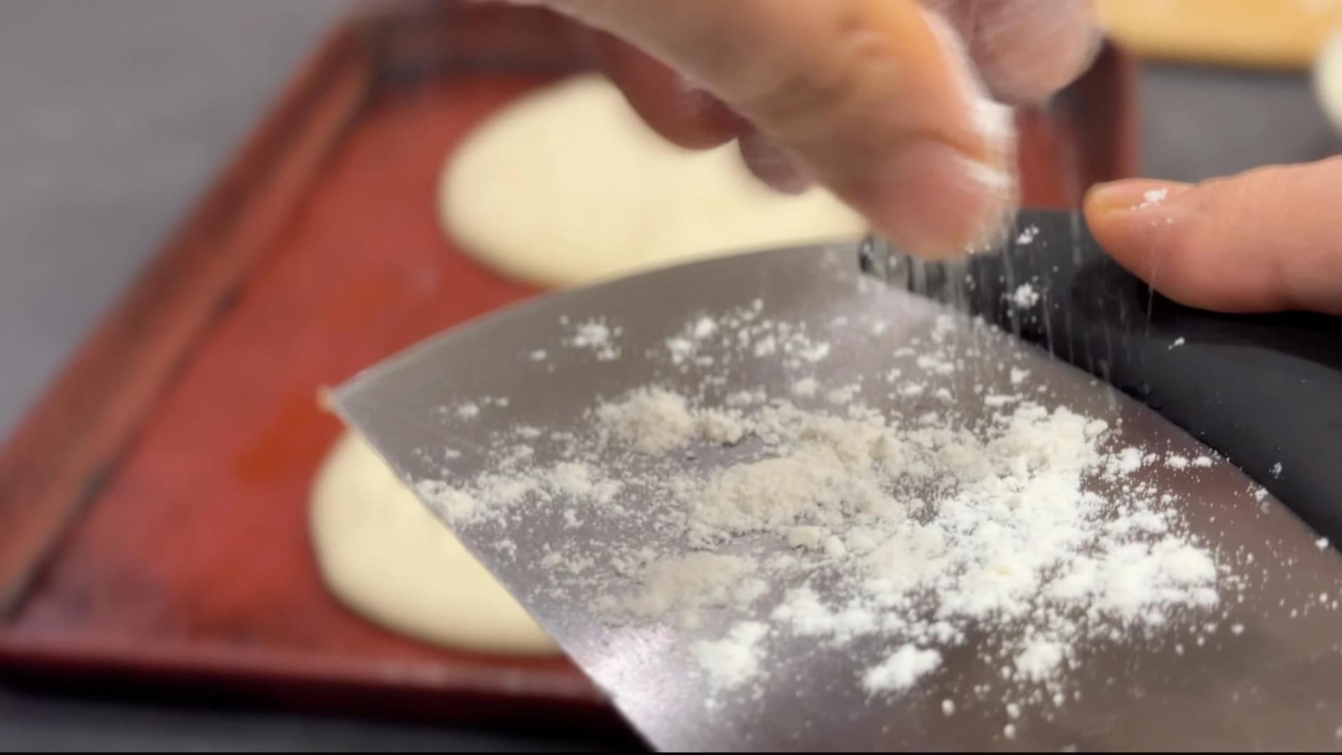 Hands dusting pizza dough with flour on a dark work surface with a red baking tray in background