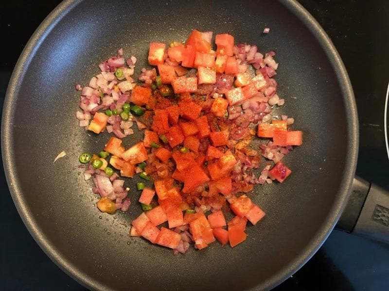 Diced tomatoes, onions, and green chilies cooking in a dark pan with spices during masala preparation.
