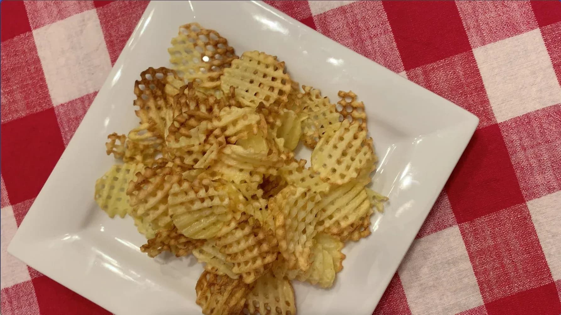 Golden crispy waffle fries piled on a white square plate over a red and white checkered tablecloth.