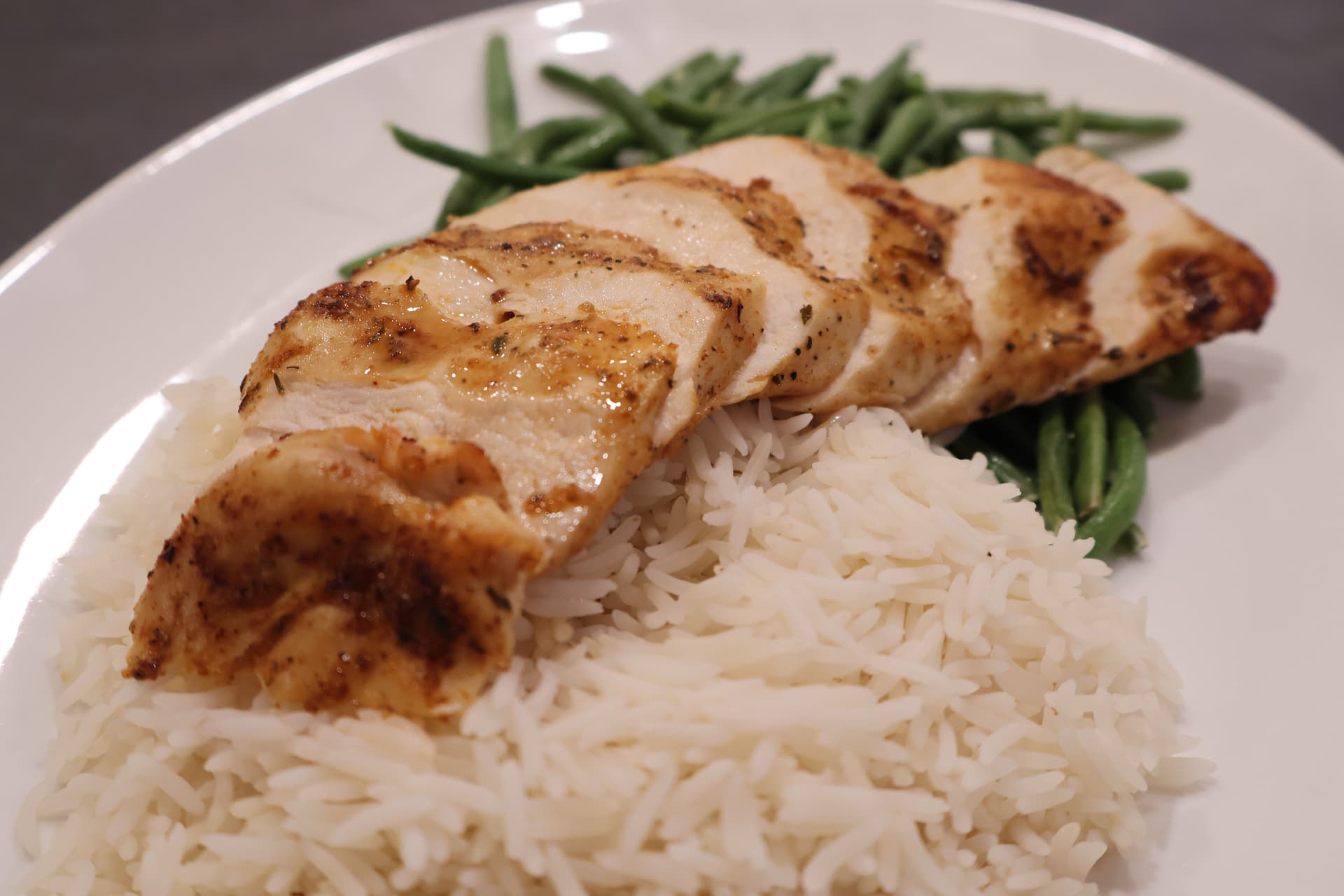 Sliced air fryer chicken breast with grill marks served over white rice and green beans on a white plate.