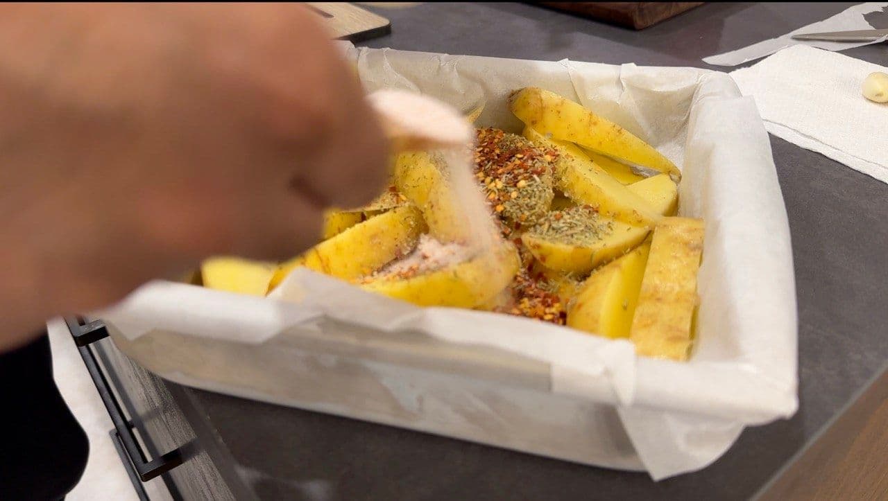 Hand seasoning golden baked potato wedges with herbs in a white takeout container.
