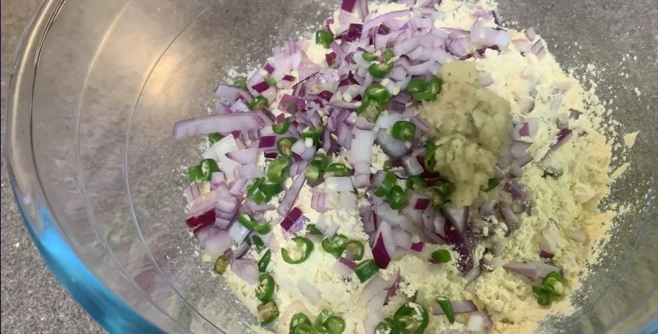 Dry ingredients for besan chilla mixed in a glass bowl: chickpea flour, diced purple onions, green chilies, and spices.