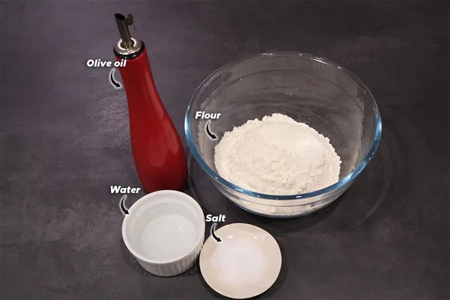 Ingredients for Flammkuchen dough: flour in glass bowl, olive oil, water, and salt on dark surface.