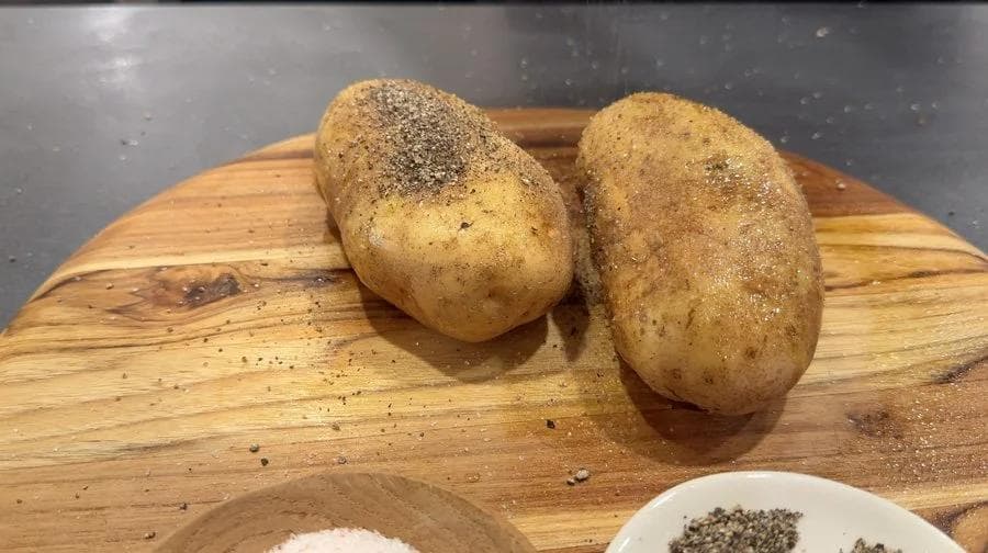 Two russet potatoes rubbed with olive oil and seasoned with salt and pepper on a wooden cutting board.
