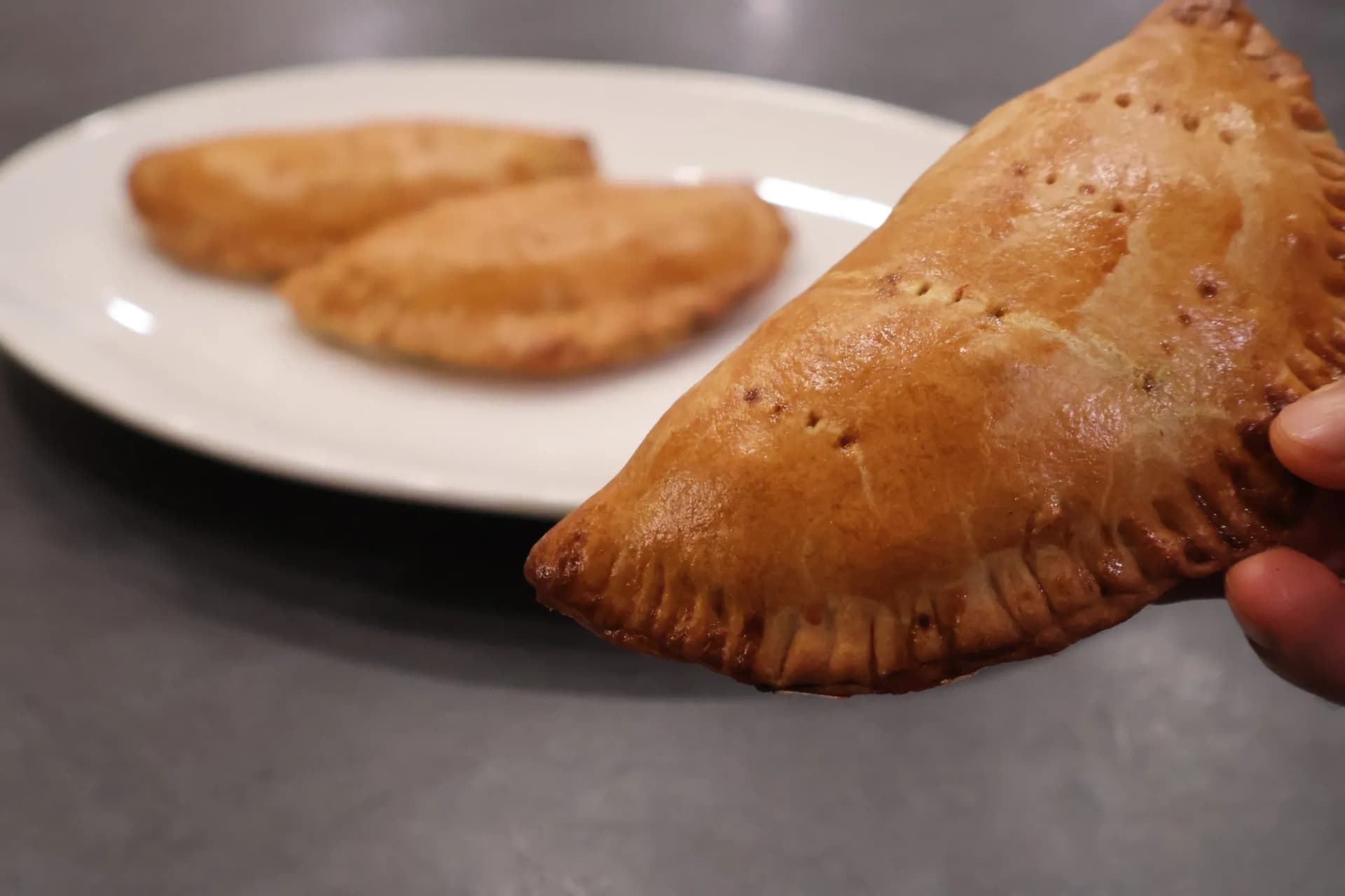 Hand holding a golden-brown Nigerian meat pie with a crimped edge, with more pies visible on a white plate in the background.