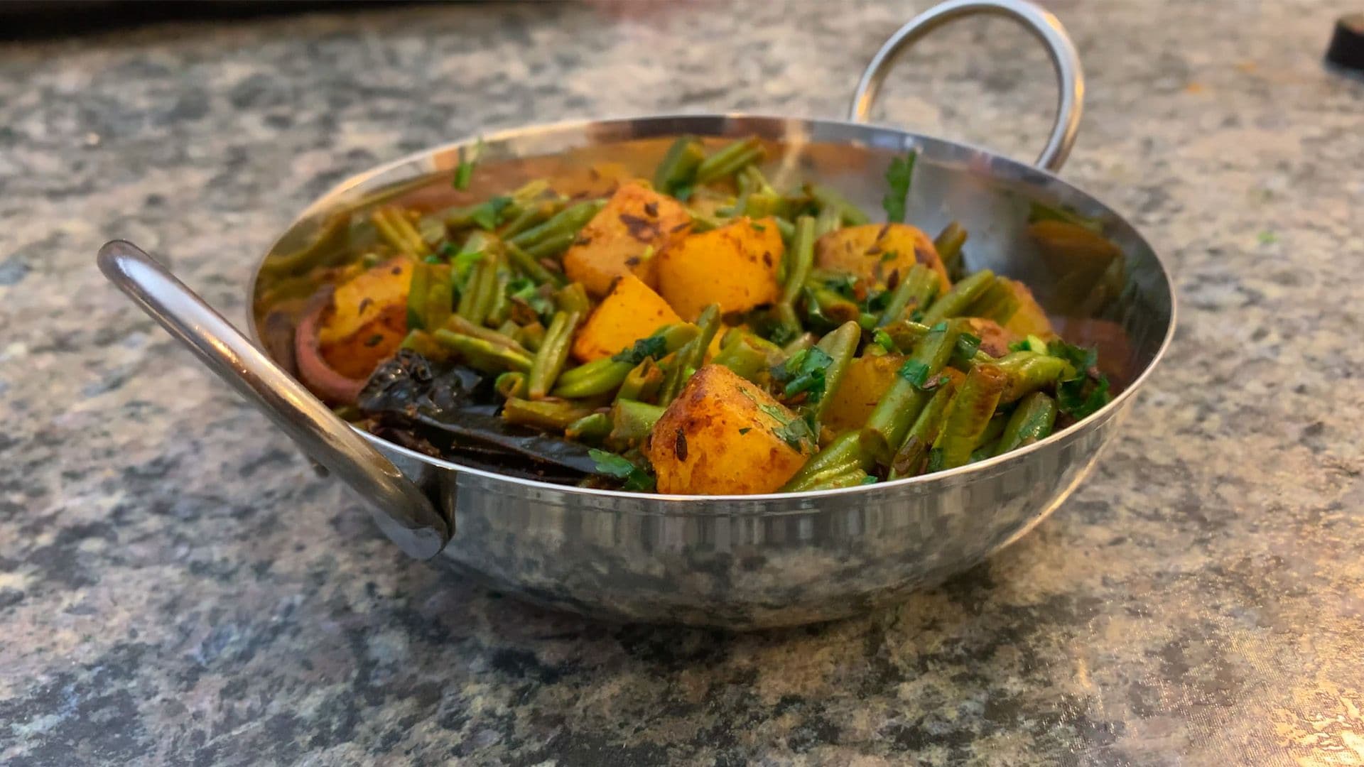 Aloo beans sabji with golden potatoes and green beans in a stainless steel wok on a stone surface.