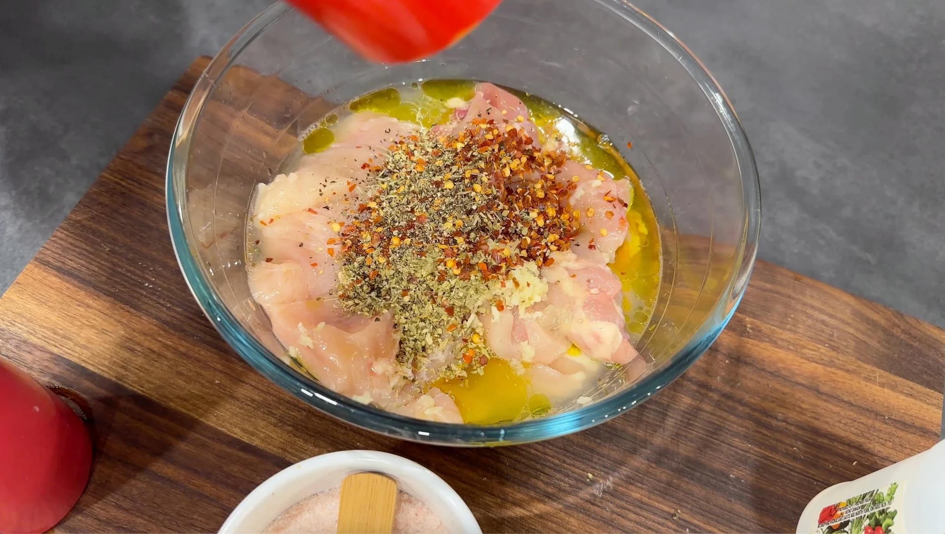 Raw chicken cubes marinating in olive oil and Greek seasonings in a glass bowl on a wooden board.