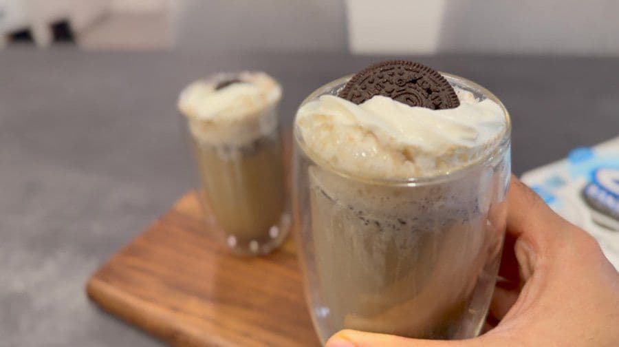 Two glasses of Cookies & Cream Oreo lattes topped with whipped cream and Oreo cookies on a wooden board.