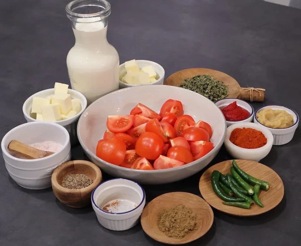 Ingredients for dal makhani laid out on a dark surface: tomatoes, butter, milk, cream, spices, green chilies, and fresh herbs in small bowls.