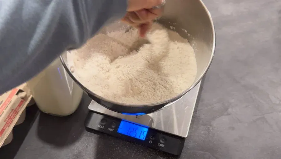 Measuring flour and dry ingredients in a white bowl on a digital scale for dinner roll dough.