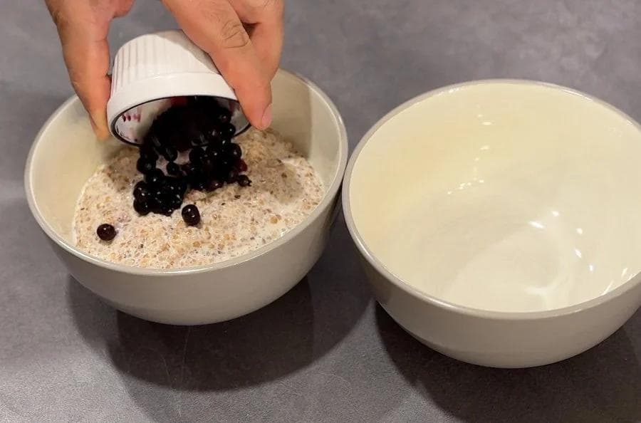 Pouring raisins and dark berries from a small cup into a bowl of cracked wheat porridge mixture.