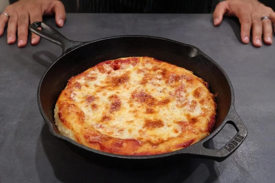 Golden-brown cast iron skillet pizza with melted cheese and crispy crust edges, held by hands over gray surface.
