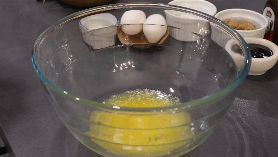 Beaten eggs and melted butter mixed with brown sugar in a glass bowl, with eggs and small bowls of ingredients nearby.