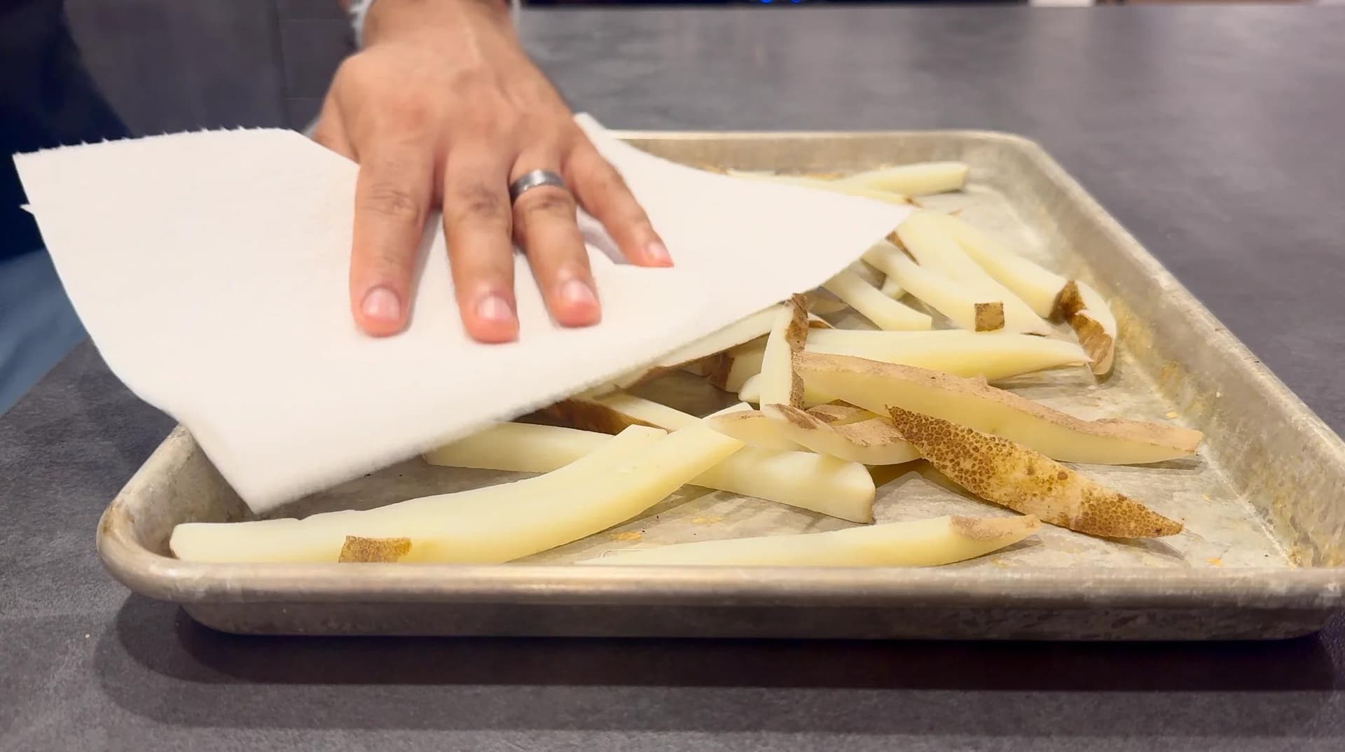 Hand patting dry boiled potato fries with paper towel on a tray before air frying.